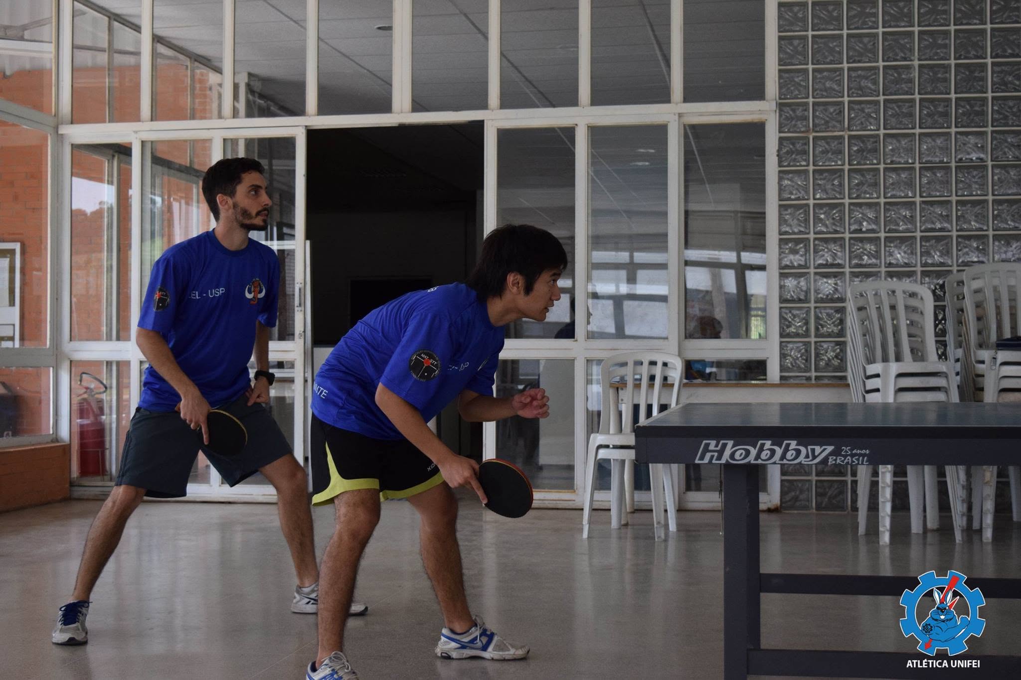 Atleta de azul inclinada junto à mesa e pessoa de laranja do outro lado.