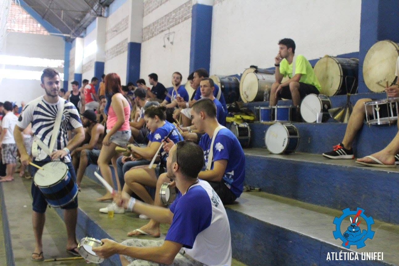 Participantes e instrumentos de percussão nas arquibancadas do ginásio; homem com tambor à frente e vários tambores grandes nos degraus.