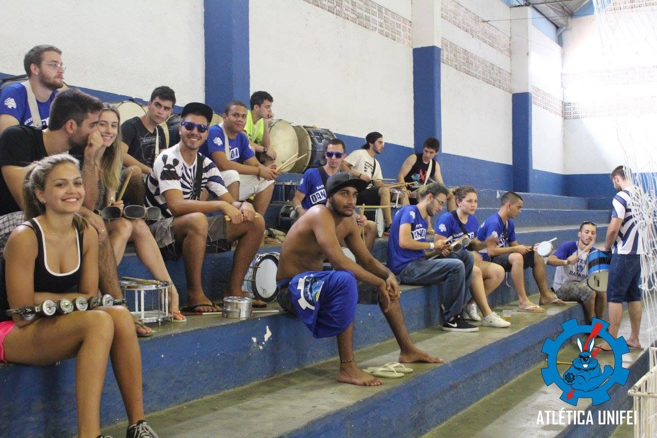 Torcedores sentados na arquibancada com instrumentos de percussão e camisetas azuis; marca Atlética UNIFEI no canto inferior.