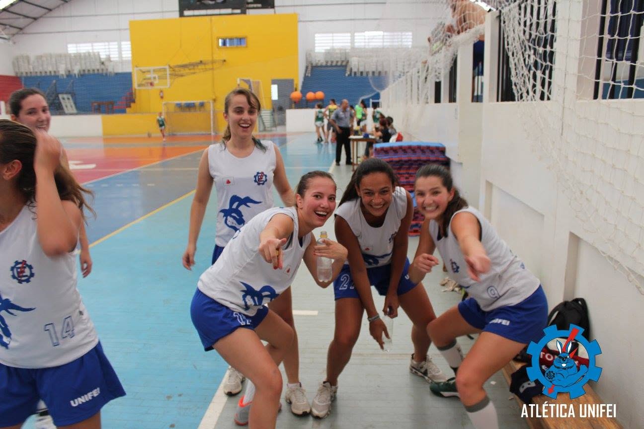 Jogadoras de uniformes brancos e azuis posam descontraídas junto à lateral da quadra; algumas apontam para a câmera.
