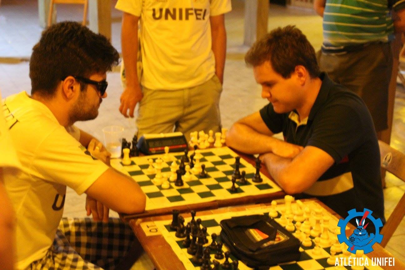 Dois homens jogam xadrez sentados à mesa, com relógio de partida e observadores; camiseta UNIFEI em participante ao fundo.