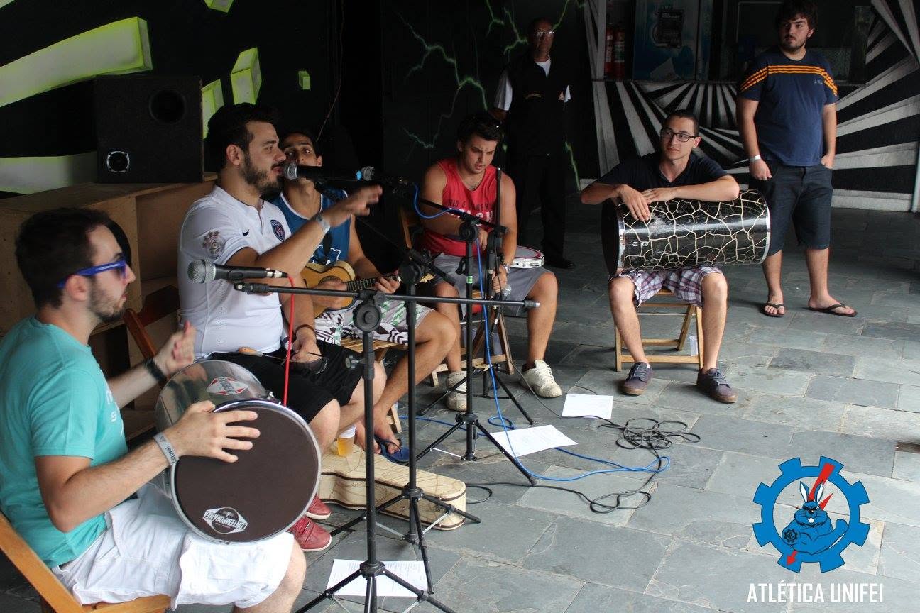Músicos sentados com instrumentos de cordas e percussão, microfones e cabos, em área coberta de paredes escuras.
