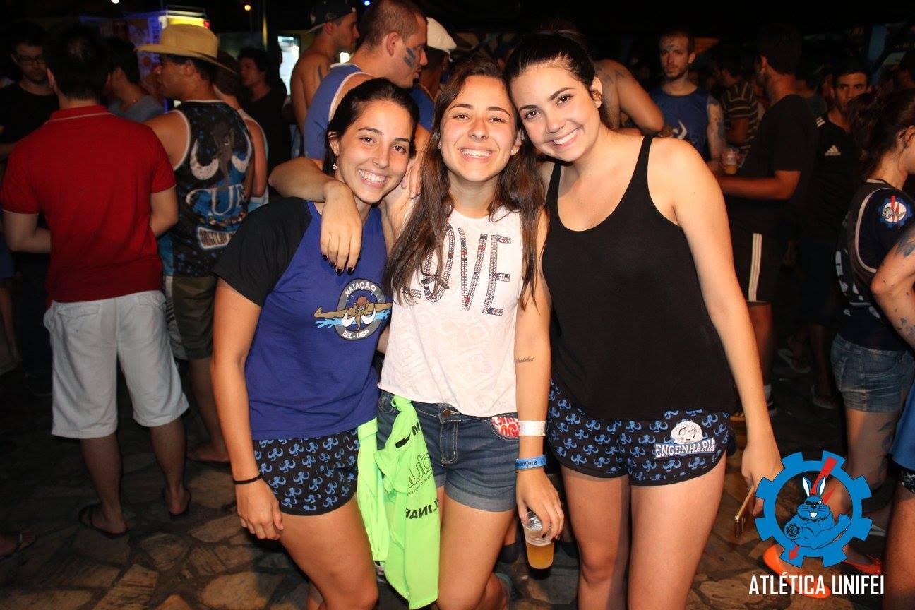 Três mulheres abraçadas posam em confraternização noturna; copos nas mãos e participantes ao fundo.